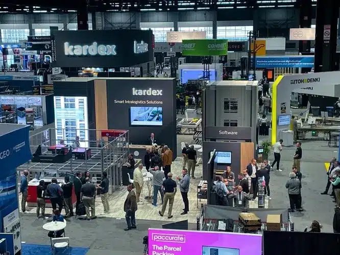 Busy warehouse automation trade show floor with multiple exhibitor booths, intralogistics solutions on display, and attendees engaging with automation and software demos
